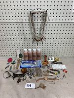 Wide view of assorted women's jewelry and fashion accessories laid out against pegboard on a table. Includes necklaces, earrings, rings, brooches, watch, makeup foundation bottles, hair clips, and small items.