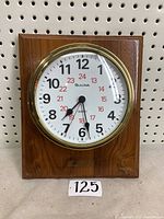 Front view of a Bulova wall mount clock with wooden frame. Shows white round clock face with black 12-hour and red 24-hour numbers, brass bezel, black hour and minute hands.