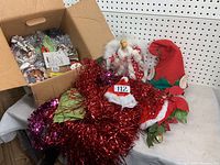 Box with assorted Christmas decorations including light-up angel, red elf hat with green leaf trim, red Santa-style hat with fur trim and bell collar, and red tinsel garland.