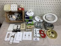 Wide view of electrical lot showing multi-plugs, outlets, switches, plate covers, boxes, and bulbs arranged on table against pegboard.