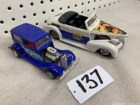 Two hot rod model cars shown from an angled top-down view, featuring a white Racing Champions 1939 Chevy with graphic on hood and a blue Majorette Hot Rod with flame decals side-by-side.