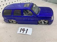 Front and side view of metallic blue 1:18 diecast Cadillac Escalade model showing closed doors and hood, chrome rims, and roof sunroof detail.