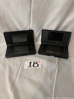 Two black Nintendo DS handheld consoles open, showing screens and controls.
