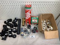 Wide shot of the plumbing lot showing multiple black ABS pipe fittings, connector pieces, a boxed Fluidmaster toilet tank repair valve, a few metal valves, and a box of white and metal toilet float hardware parts.