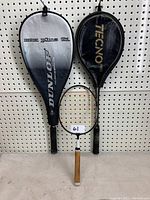 Two tennis racquets hanging with covers, one Dunlop Max Plus Ti and one Tecnopro Pro.