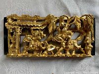 Full front view showing intricate gilt carving of warriors on horses and architectural elements in a deep relief style mounted on a wooden panel.