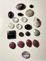 Full set of 18 polished cabochon gemstones shown on white background, variety of colors and shapes visible.