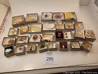 Top-down view of all 28 Avon jewelry pieces in grey striped boxes, showing variety of pins, brooches, lockets, and earrings mostly in gold-tone metal with colorful and pearl embellishments.