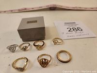 Seven rings arranged in front of an Avon box and item tag, showing variety of styles and sizes.