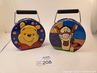 Front view of two circular embossed metal lunchboxes, one with Winnie the Pooh face and the other with Tigger face, both with black handles.