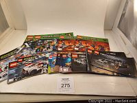 Lot of sixteen LEGO instruction manuals from multiple series displayed open and stacked flat on a white backdrop.