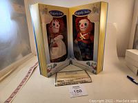 Wide photo showing both Raggedy Ann and Raggedy Andy dolls in open display box with certificate.