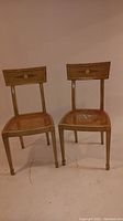 Two light cream painted wooden chairs with decorative gold and floral accents on the backrest. Cane seats visible with damage.