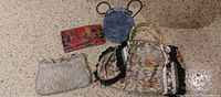 Photo showing four ladies handbags on the floor: cream textured handbag, small round blue handbag with chain, red-black-gold patterned clutch, large multicolor paisley handbag with gold handles