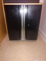 Front view of the Husky 2-door steel base cabinet showing black doors with silver vertical handles and light wood-colored top, placed on castors.