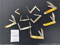 Nine vintage pocket tools arranged on black cloth, showing multiple knives with varying handle colors and blade sizes and shapes.