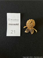 Top view of the gold-toned blooming flower brooch next to a MaxSold label number 21 on a black background.