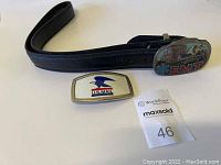 Photo showing black leather belt with attached colorful EMT belt buckle and separate USPS belt buckle.