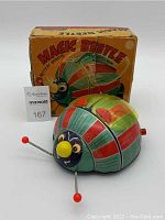 Photo of the vintage Magic Beetle tin litho toy with its colorful striped body, yellow nose, and red-tipped metal antennae in front of the matching original box.