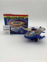 Photo of the vintage wind-up space ship toy with the original box behind it, showing the colorful rotor and cockpit details.