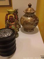 Oriental ladder jar, smiling Buddha statue holding a gold ingot, and brass candle holders displayed on a surface