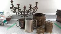 Full lot showing five-arm brass candleholder, brass cup, tankards, and containers on countertop with green tape markers.