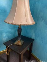 Photo showing the full lamp with brass base and cream fabric shade positioned on the dark wood side table against a blue wall.