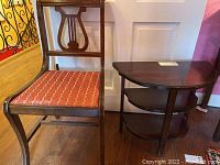 Photo of wood side chair with lyre back and reddish-orange upholstered seat next to a two-shelf wood side table with scratches.