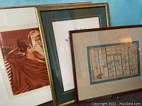 Three framed artworks stacked leaning against a wall, showing partial view of each.