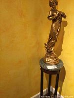Full view of the gold colour lady statue on marble-topped wooden stand in corner against yellow wall.