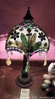 Full view of lamp showing colorful fabric shade with green and brown leaf design, multicolored beads around the edge and hanging glass droplet pendants. Ornate cast metal base with green glass accent visible.