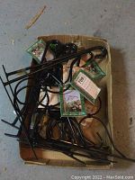 Photo showing ten black metal plant brackets piled in a box with tags attached.
