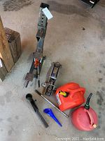 Photo showing automotive hydraulic floor jack, tall bottle jack with height holes, red plastic gas can, red metal gas container, two blue funnels, and metal jack handle extension bar on concrete floor.
