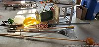 Photo showing assortment of gardening tools, bird feeder box, seed spreader, and other garden accessories arranged on floor with sunlight.