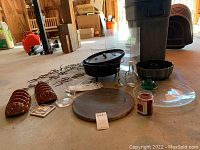 Full view of all items in lot arranged on the floor. Includes cast iron Dutch oven, pizza stone, ceramic taco holders, glass tray, candle, and other kitchenware items.