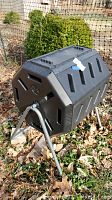 Black plastic tumbler composting bin on a metal A-frame stand in an outdoor area with fallen leaves.