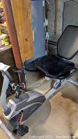 Recumbent stationary exercise bike in a basement setting showing frame, pedals, and cushioned seat with cover.