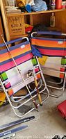 Two multi-colored striped folding beach chairs with aluminum frames and blue padded armrest covers, leaning against a wooden shelf. True Temper fishing pole with wooden handle and Compac 6000 reel placed around the chairs.