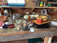 Full view of kitchen items on wooden table including various pots, glassware, pottery bowls, baskets, and cookie cutter