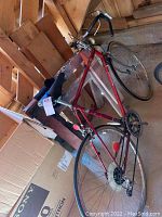 Side view of red Panasonic Sport 1000 bike leaning against a wooden surface and cardboard box, showing frame, wheels, handlebars, and saddle.
