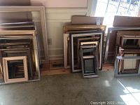 Photo showing multiple picture frames stacked against a corner wall with various sizes and finishes including wood and gold tones.