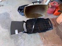 Image showing the open portable Weber grill, hinged beige lid, black cooking grate, side handle, and black storage bag beneath.