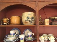 Shelving unit with multiple items: stoneware crock with cobalt floral decoration, brown drip glaze pot, Studio Nova dishes, yellow lidded tureen, decorative globe and vintage scale