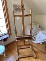 Full frontal image of wooden standing art easel showing height and structure near window with wood flooring and wall art in background.