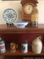 Photo shows decorative blue and white plate, matching bowl, stoneware whiskey jug, winding wooden clock, and three different beer steins (silver, brown, gray).