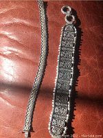 Two sterling silver bracelets placed side by side on a leather surface: narrow linked chain bracelet and wide patterned filigree bracelet.