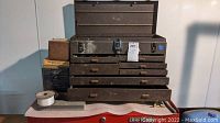Closed view of large brown metal toolbox with multiple drawers, several small boxes stacked to the side. Shows surface wear, oxidation, signs of use.