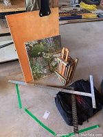 Wooden French easel, floral canvas painting, metal T-square, angle ruler, and black easel bag arranged on concrete floor showing wear and staining.