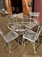 Photo showing white wrought iron patio set with one round metal table base (no glass top) and four chairs arranged outdoors with a barn and grass background.