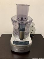 Front view of food processor showing silver base with control buttons and clear plastic bowl and lid with feed tube.